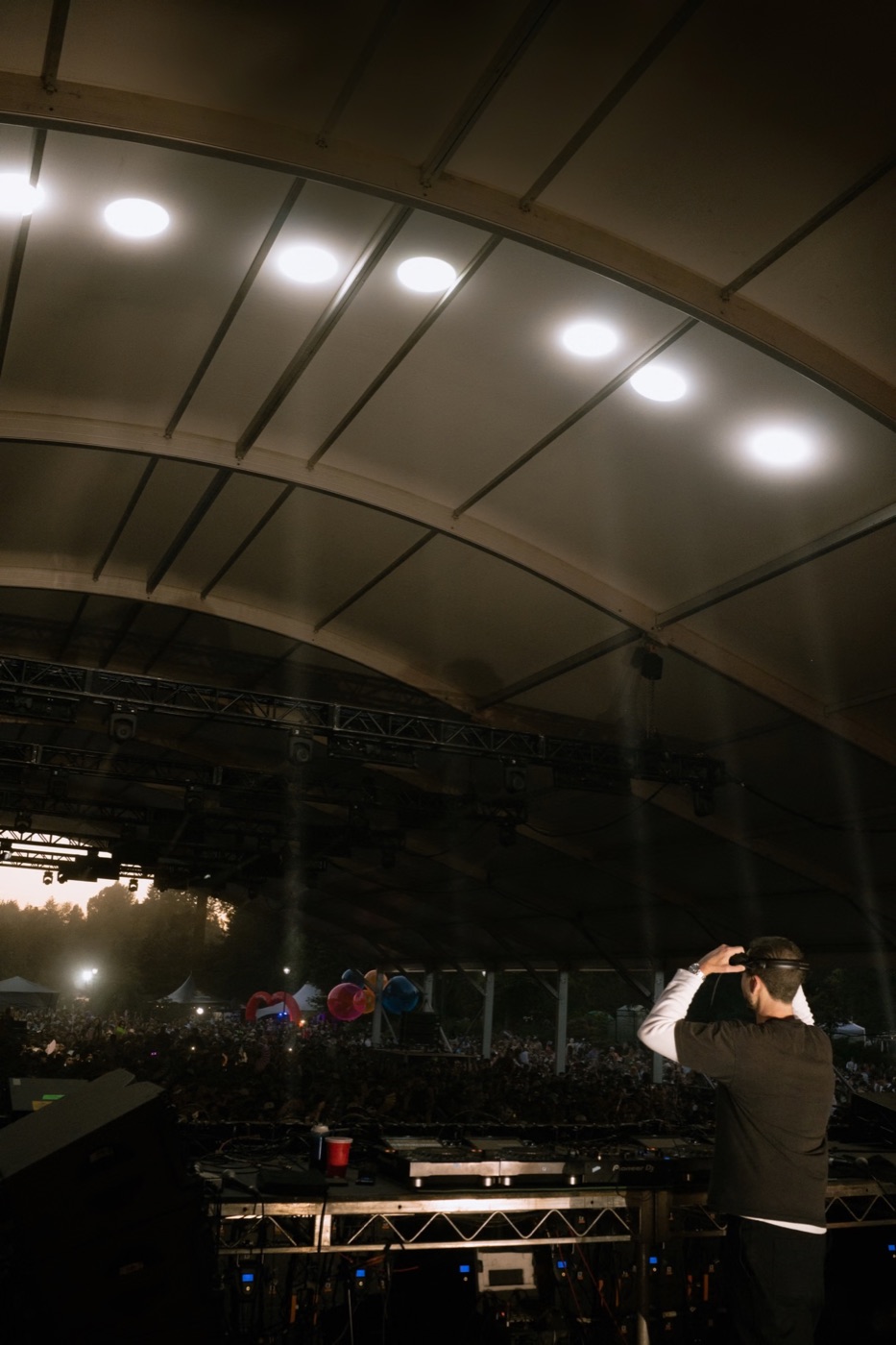 a dj adjusting headphones under a lit canopy, looking out over the crowd at dusk