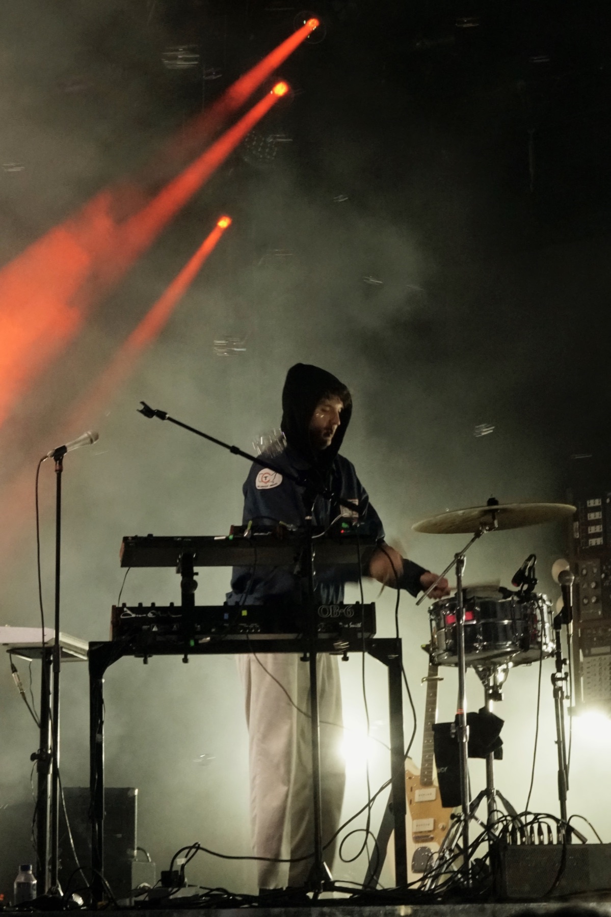 a hooded musician playing synths and a snare under red stage lights and haze