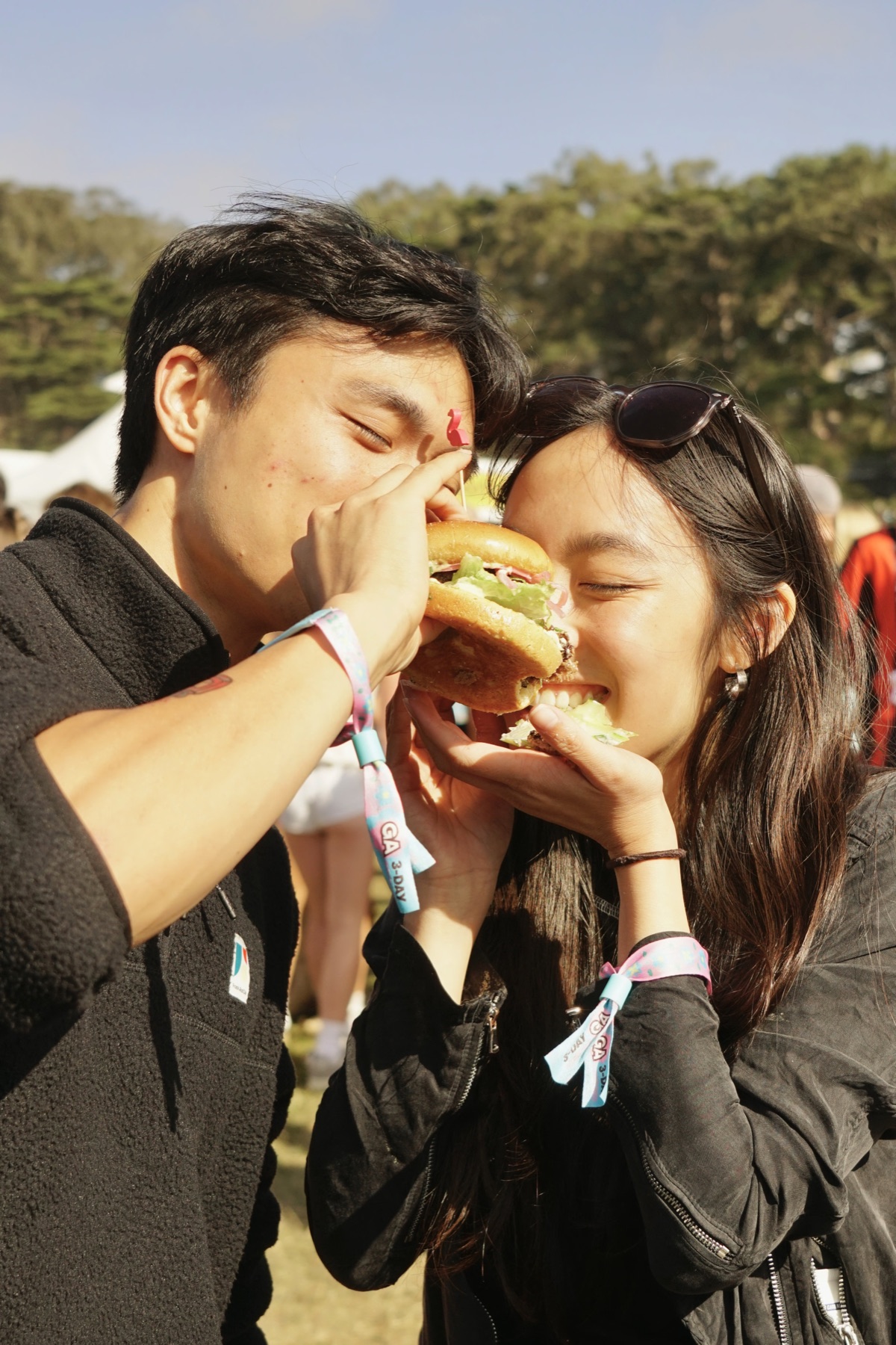 two friends sharing a bite of one burger at a festival
